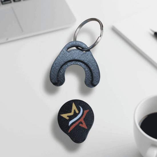 A black keychain and guitar pick on a white desk with a laptop and coffee cup.